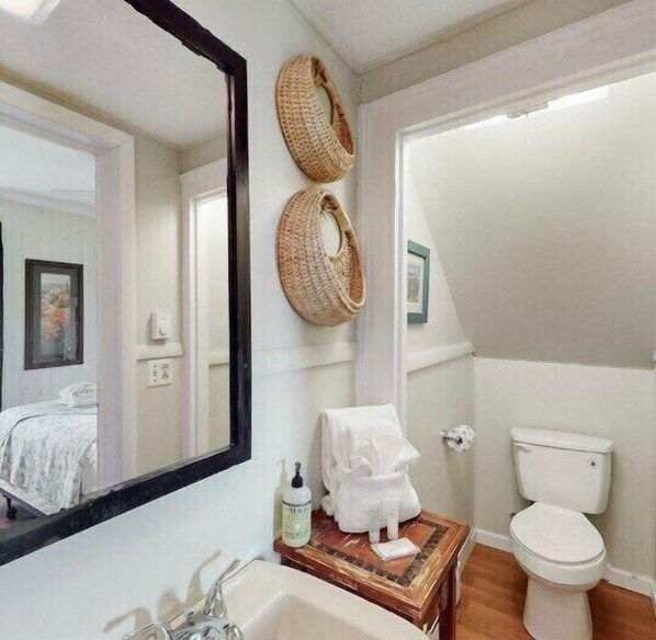 Un baño con un espejo grande, dos cestas tejidas, un lavabo con artículos de aseo, un inodoro y un cuadro enmarcado en la pared.