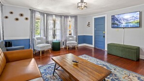 A living room with a wooden coffee table, two patterned armchairs, a green ottoman, a blue door, and a TV on the wall. - Walk to IthacaCommons+Bars+Shops (Ithaca)