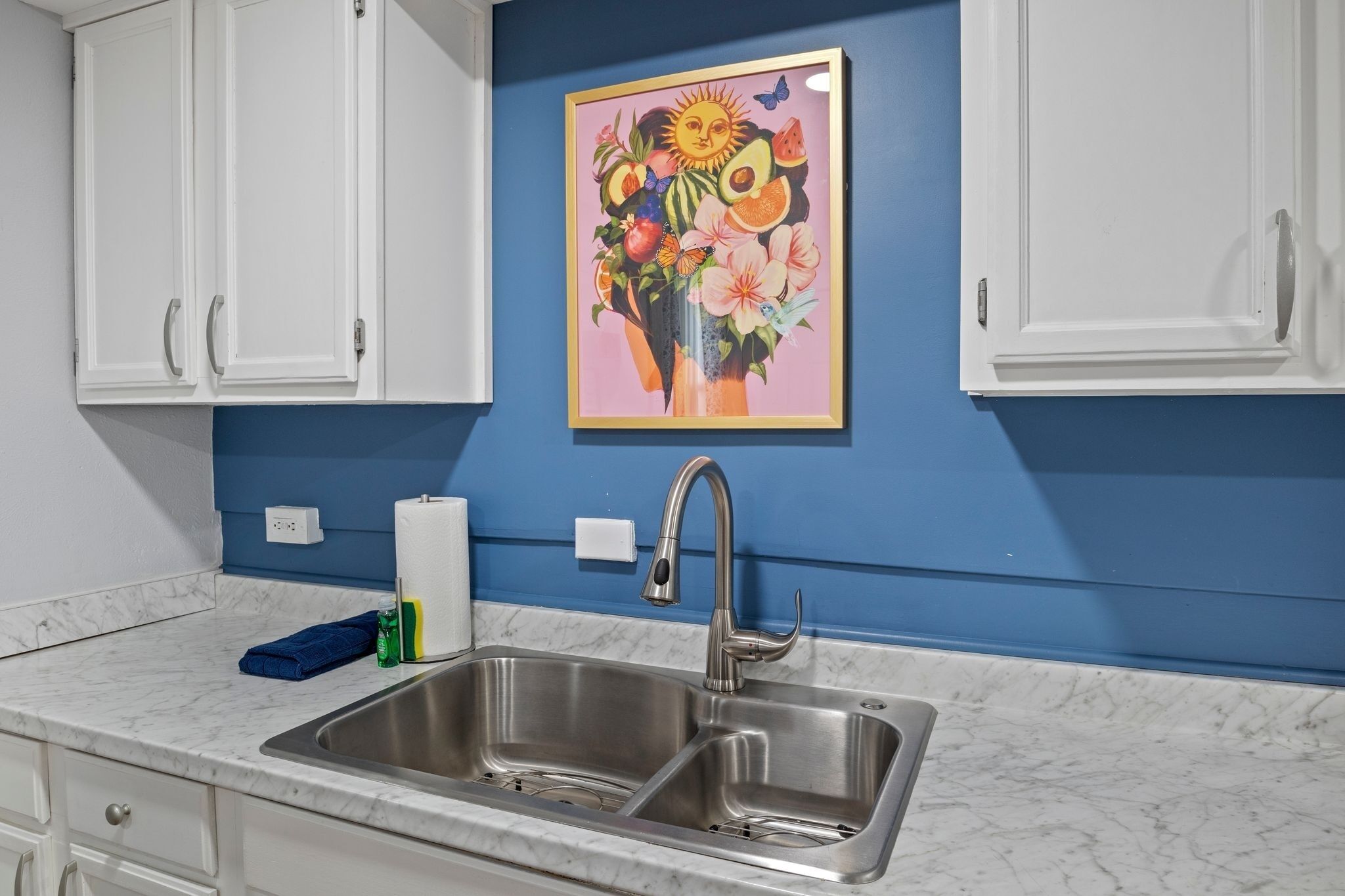 Een keuken met een dubbele spoelbak, witte kastjes, een blauwe wand en een kleurrijk schilderij boven de spoelbak.