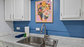 Een keuken met een dubbele spoelbak, witte kastjes, een blauwe wand en een kleurrijk schilderij boven de spoelbak.