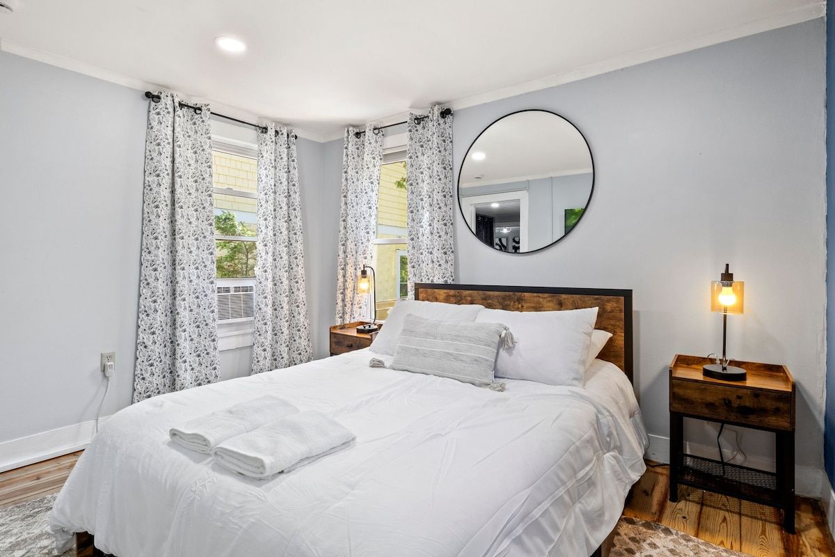 Quarto com cama grande, espelho redondo, abajur e janela com cortinas.