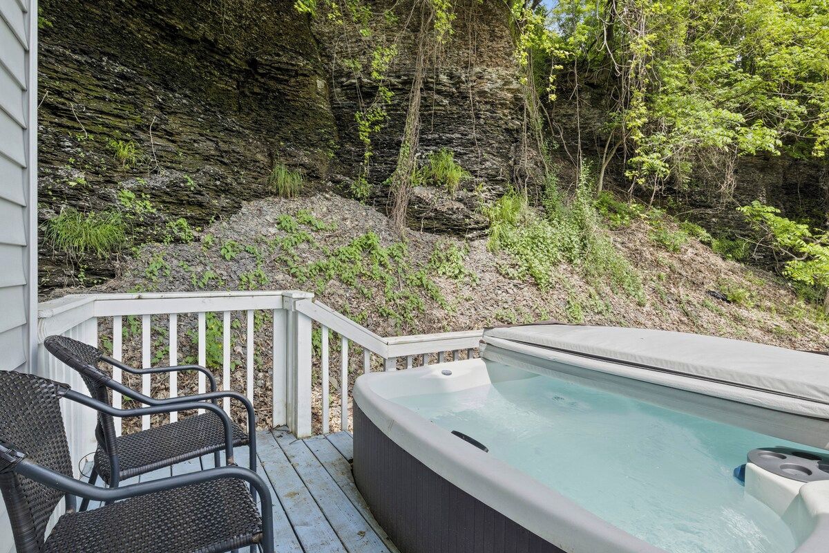 Un jacuzzi en una terraza de madera, con un risco rocoso y árboles al fondo.