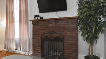 Una sala de estar con chimenea de ladrillo, un televisor montado encima y una planta en maceta a la derecha.