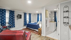 Une chambre à coucher comprenant un lit, un fauteuil rouge, une petite table, un téléviseur, et une salle de bain avec un miroir et un porte-serviettes.