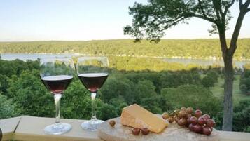 Zwei Gläser Rotwein, ein Käsekeil und Trauben auf einem Holzboden, im Hintergrund ein See und ein Wald.
