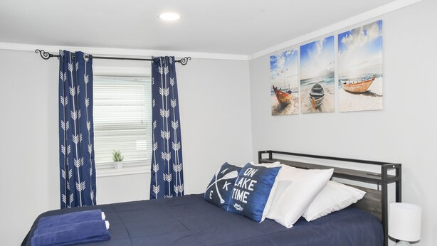 A neatly made bed with a dark blue bedspread and matching pillows, a window with white blinds, and a wall with three framed pictures of boats on a beach.