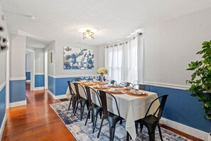 Un comedor con una mesa larga dispuesta para seis personas, sillas, un florero con flores y un cuadro en la pared.