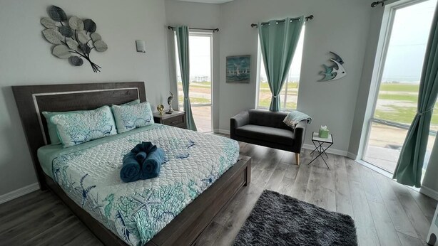 A bedroom with a bed, a chair, a rug, and a view of the outdoors. - Beachy Escape for 2 Fun in The Sun Endless Charm! (Galveston)