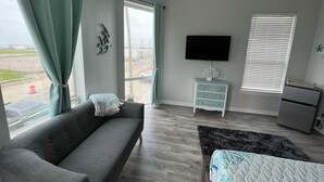 A modern bedroom with a grey sofa, a bed with a patterned bedspread, a television on the wall, and a window with blinds. - Beachy Escape for 2 Fun in The Sun Endless Charm! (Galveston)