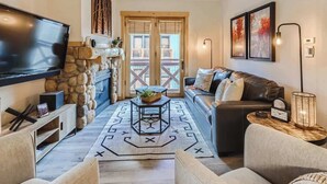 A cozy living room with a stone fireplace, a large flat-screen TV, a leather sofa, and a dining table with a geometric rug. - Luxury Slope & Village Views, Deck, Steps to Lifts (Frisco)