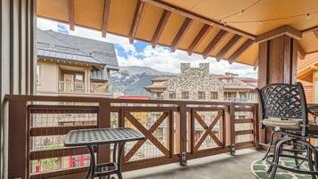A balcony with a table and chairs, a view of buildings and mountains, and a wooden ceiling structure.