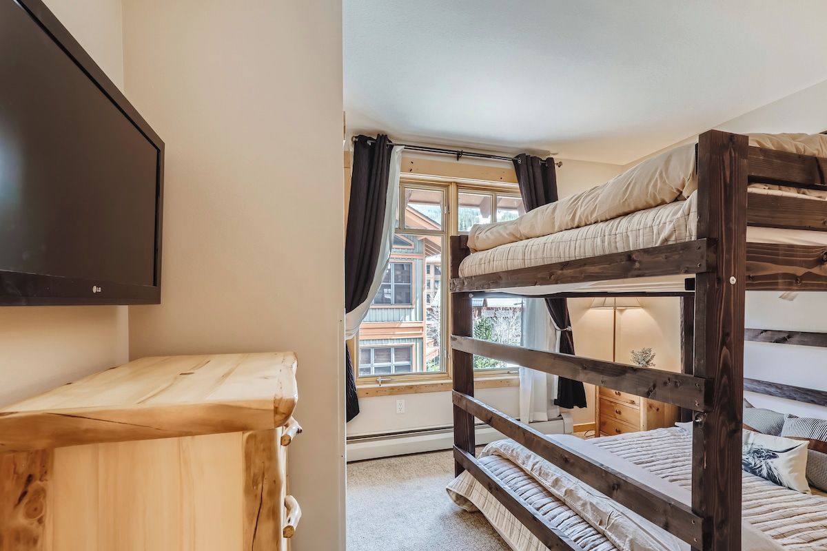 A bunk bed room with a TV on the wall, a wooden dresser, and a window with curtains.