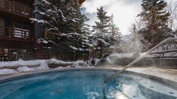 Un bain à remous entouré d’arbres couverts de neige et un bâtiment en bois doté de balcons.