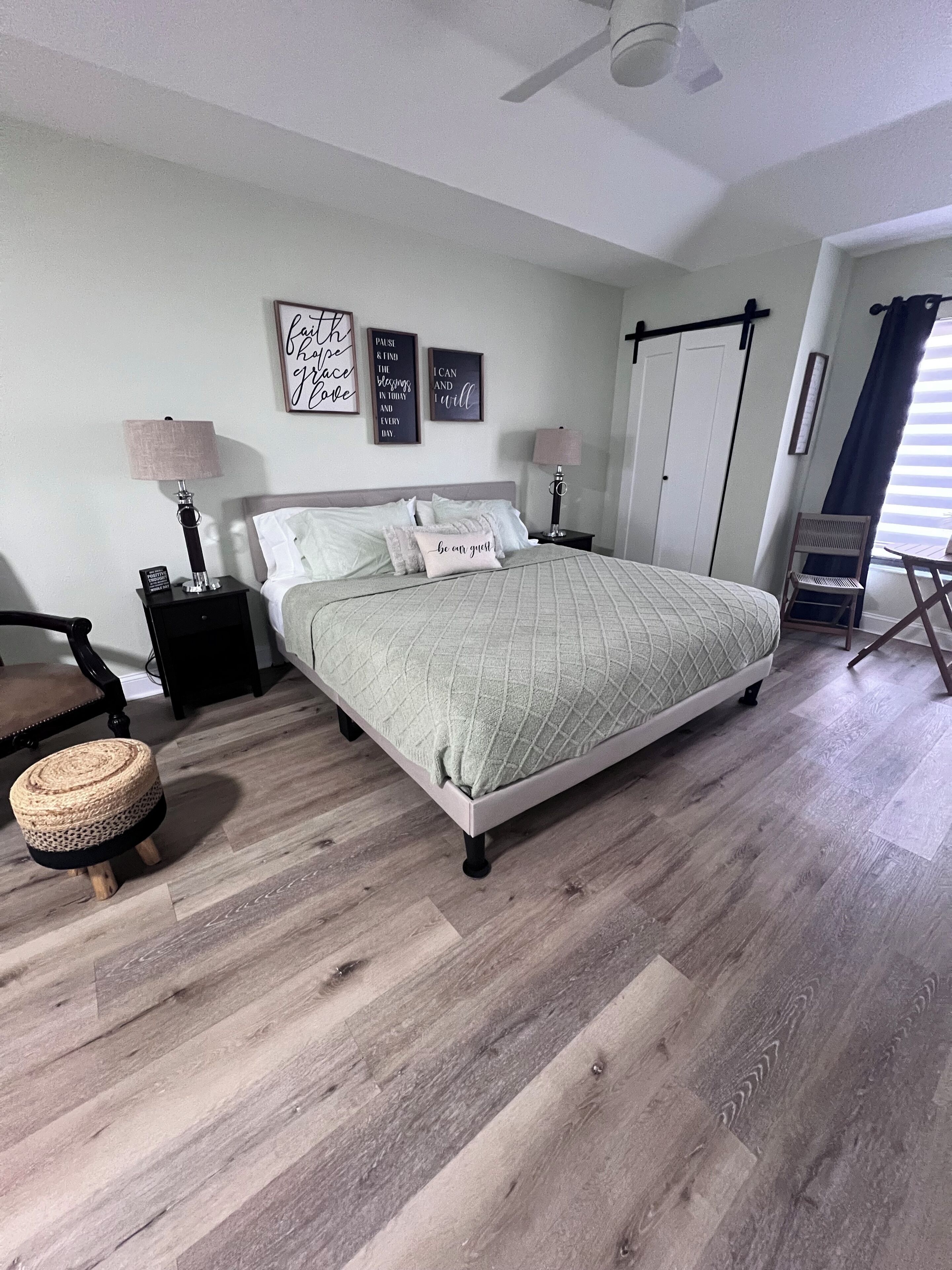 Un dormitorio ordenado con una cama, mesitas de noche, una silla y un taburete pequeño.
