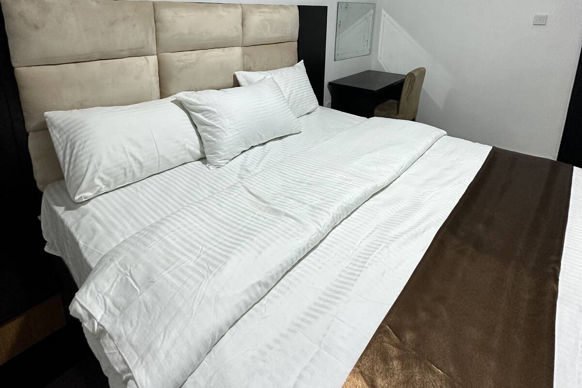 Un lit bien fait, avec du linge de lit blanc et un élément décoratif marron, une table de chevet et un petit miroir accroché au mur.