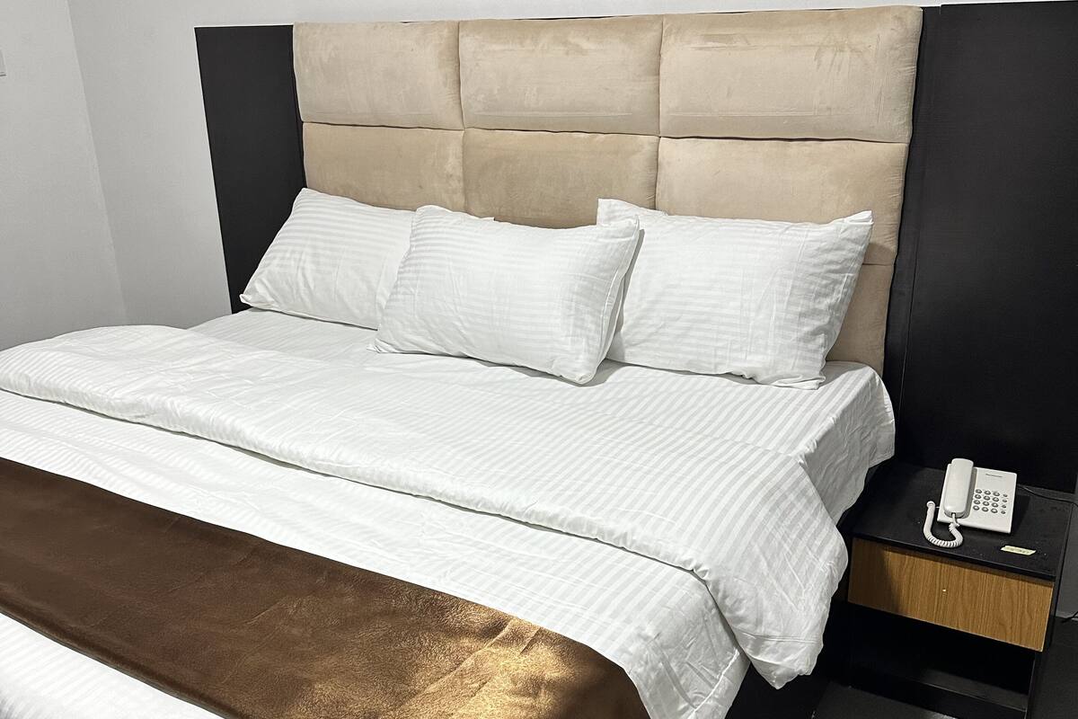 Un lit bien fait, avec du linge de lit blanc et une couverture marron, un téléphone sur la table de chevet et une table de nuit noire.