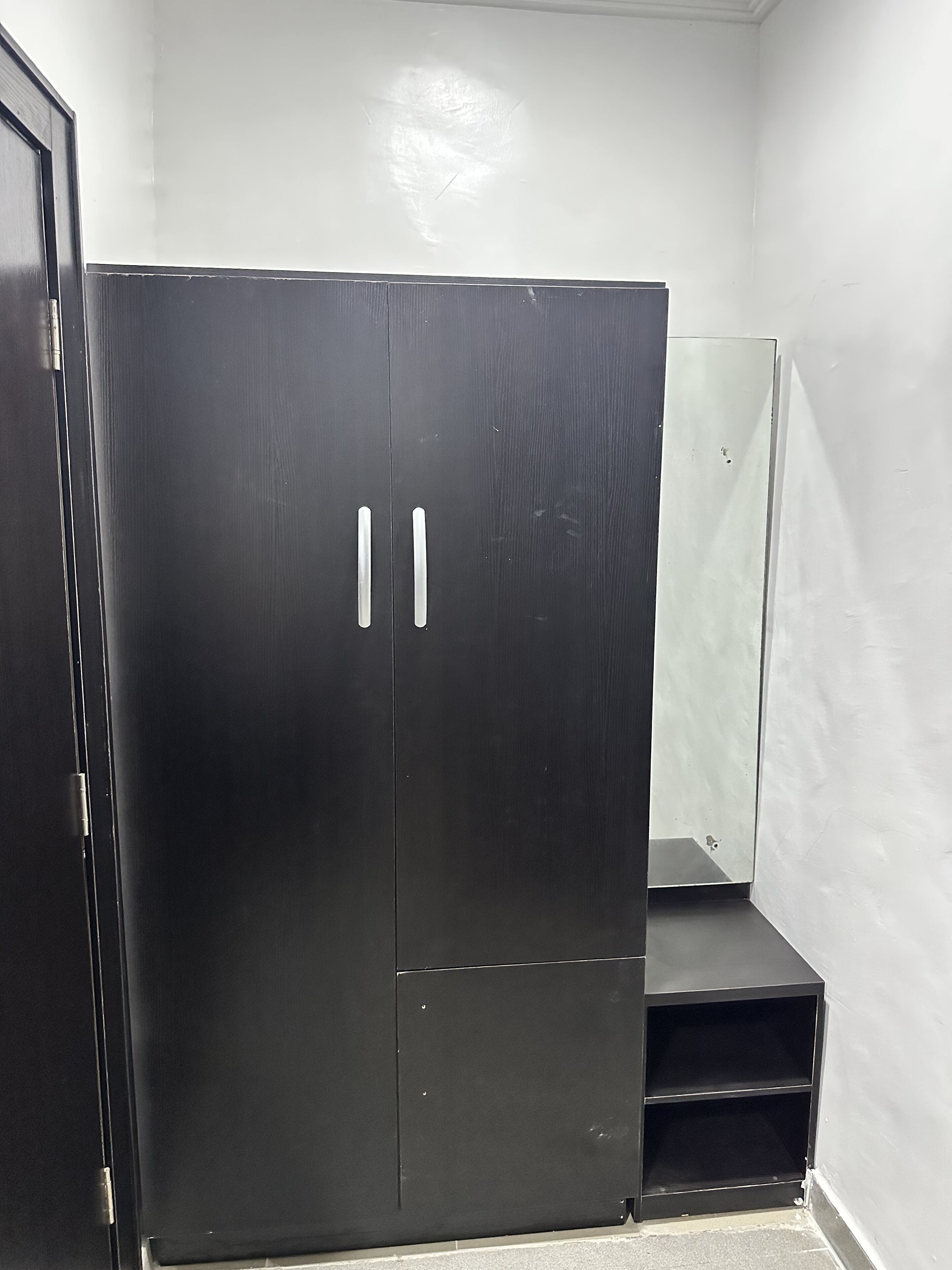 A black wardrobe with two doors and a shelf, located in a corner of a room with white walls.