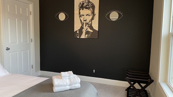 Un dormitorio con una cama, un banco tapizado gris, una puerta blanca y dos decoraciones en forma de ojo montadas en la pared.