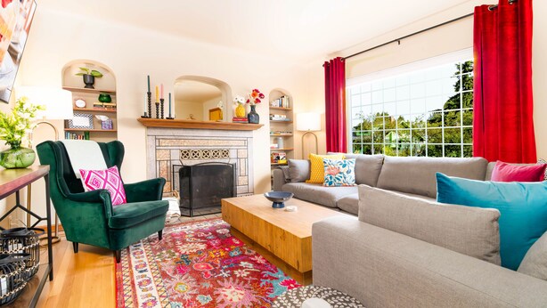 A living room with a fireplace, green armchair, grey sofa, and a colorful rug.