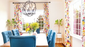 A dining area with a wooden table, blue upholstered chairs, a chandelier, and floral curtains.