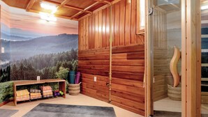 A room with wooden walls, a bench with folded blankets, a small shelf with pillows, and a large window with a view of a forest and mountains.