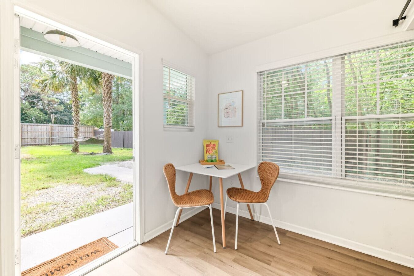 A room with a white door leading to a backyard, a small table with two wicker chairs, a framed picture on the wall, and a window with blinds.