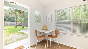 A room with a white door leading to a backyard, a small table with two wicker chairs, a framed picture on the wall, and a window with blinds.