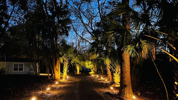 Un sentier de terre bordé d’arbres et éclairé par de petites lumières, qui mène à un bâtiment doté d’une fenêtre.