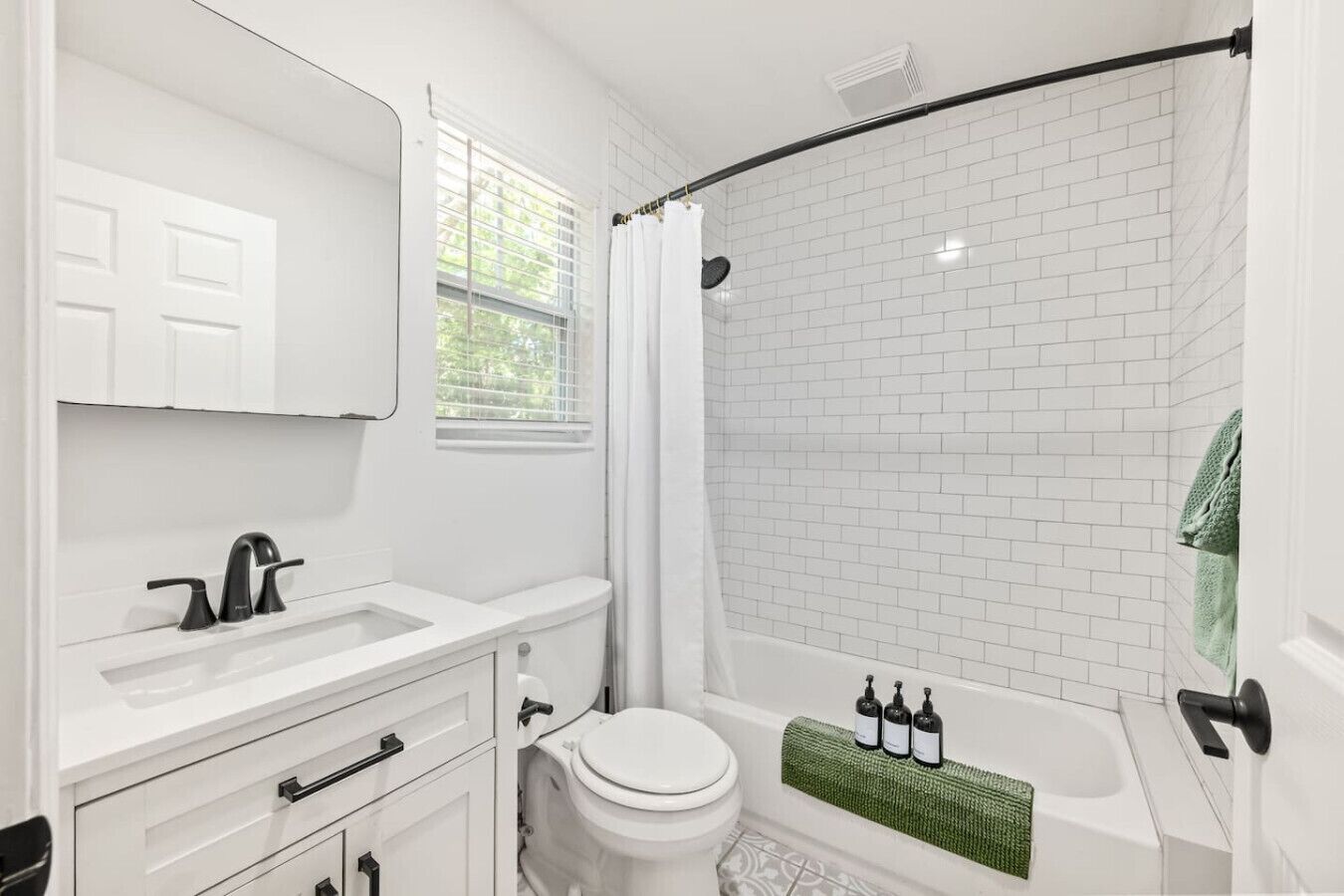 A bathroom with white fixtures, a shower with a curved door, and a window with blinds.
