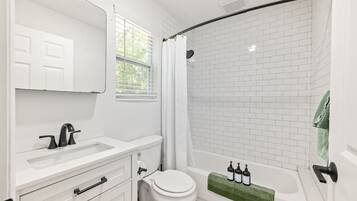 A bathroom with white fixtures, a shower with a curved door, and a window with blinds.