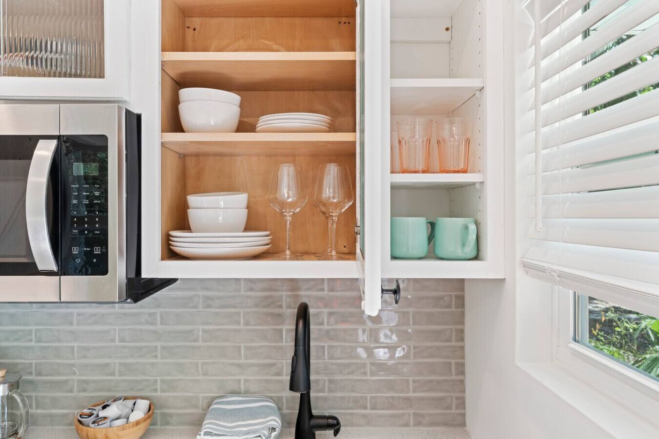 A kitchen with a microwave, a cabinet with dishes and glasses, and a window with blinds.