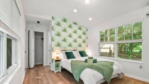 Une chambre à coucher comprenant un lit, des tables de chevet, une commode, une fenêtre donnant sur des arbres et une porte menant à une autre pièce.