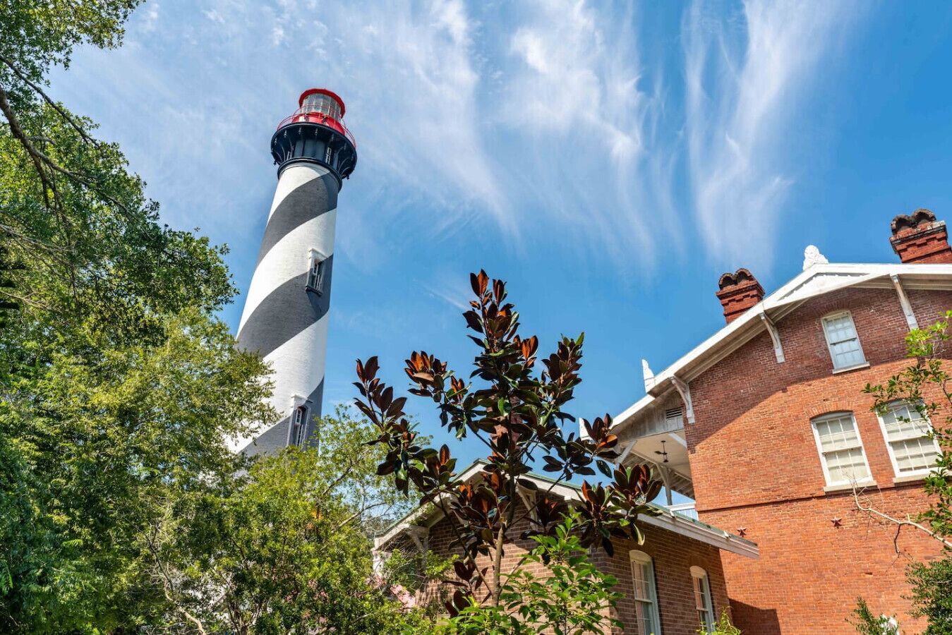 A lighthouse with a spiral design, a brick building with white windows, and surrounding greenery.