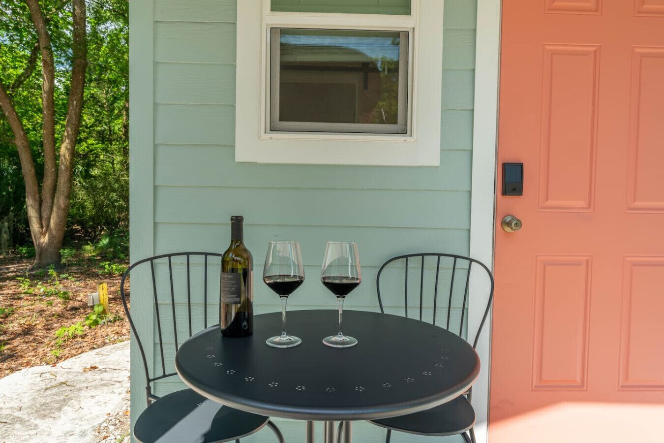 A round table with two chairs, a bottle of wine, and two wine glasses on a porch with a light blue wall and a pink door.