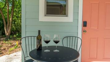 A round table with two chairs, a bottle of wine, and two wine glasses on a porch with a light blue wall and a pink door.