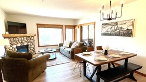 A spacious living room with a stone fireplace, a sofa, a dining table set for four, and large windows. - Feel at Home after a Day of Adventures in this Family friendly Retreat! (Canmore)