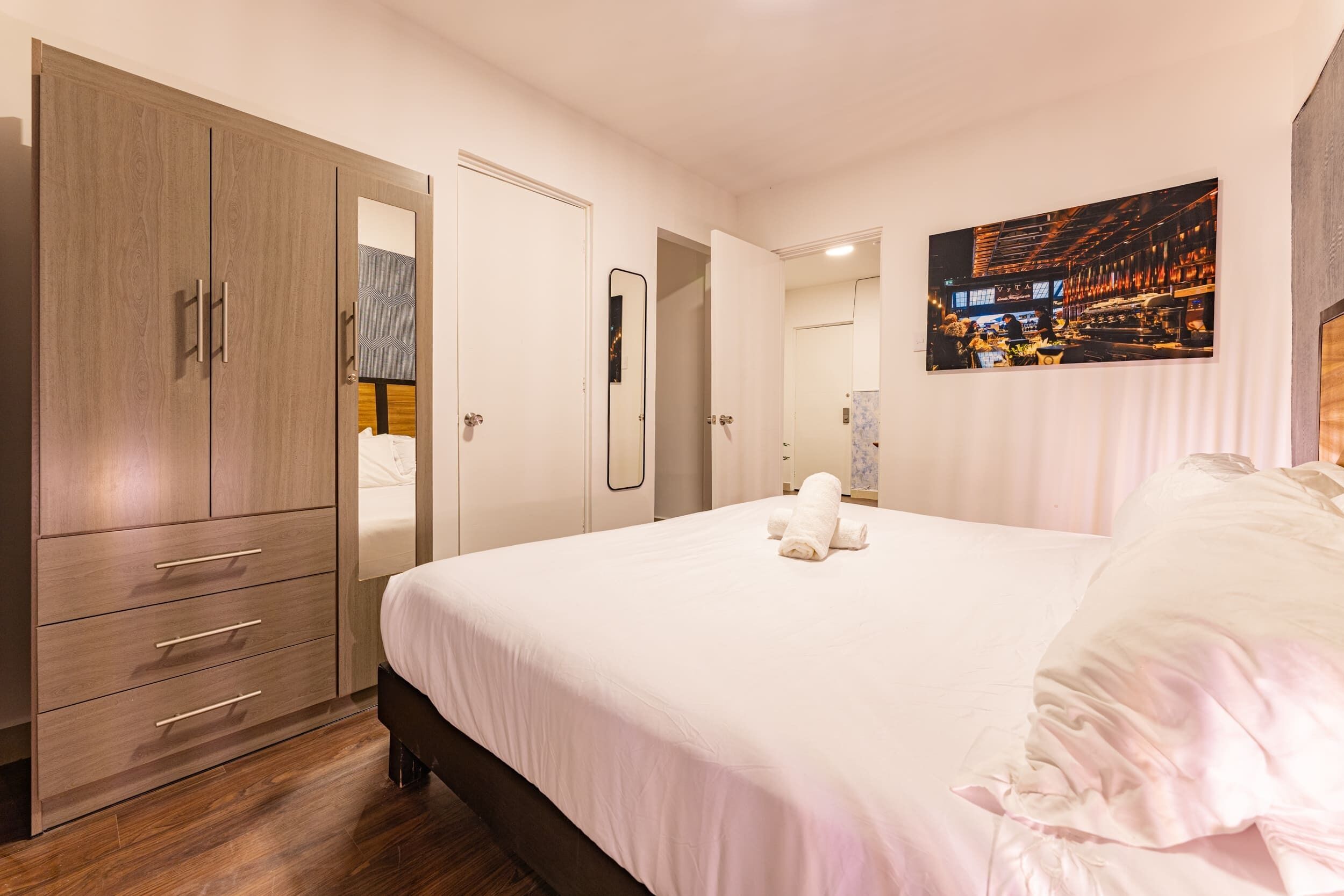 A hotel room with a large wardrobe, a bed with white bedding, a mirror, and a wall-mounted artwork.