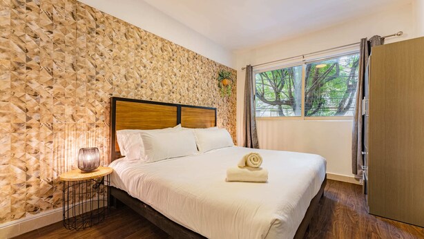 A neatly made bed with a wooden headboard, a bedside table with a lamp, and a window with curtains. - Condesa Zamora Lofts by VH (Mexico City)