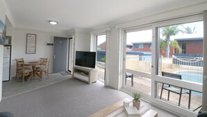 A living room with a TV, dining table, chairs, and a view of a swimming pool. - Poolside - Ground floor opposite pool (Merimbula)