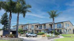A multi-story residential building with a central staircase, a covered driveway, and a landscaped front yard with palm trees and shrubs. - Poolside - Ground floor opposite pool (Merimbula)