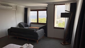 Habitación de hotel con un sofá gris, una pequeña mesa de madera, una lámpara y vistas a un paisaje urbano a través de grandes ventanales.