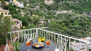 Mesa redonda com cadeiras brancas num terraço, com vista para uma paisagem montanhosa com edifícios.
