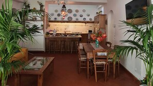 Uma sala de jantar acolhedora com cozinha ao fundo, apresentando painel de azulejos, mobiliário de madeira e plantas em vasos.