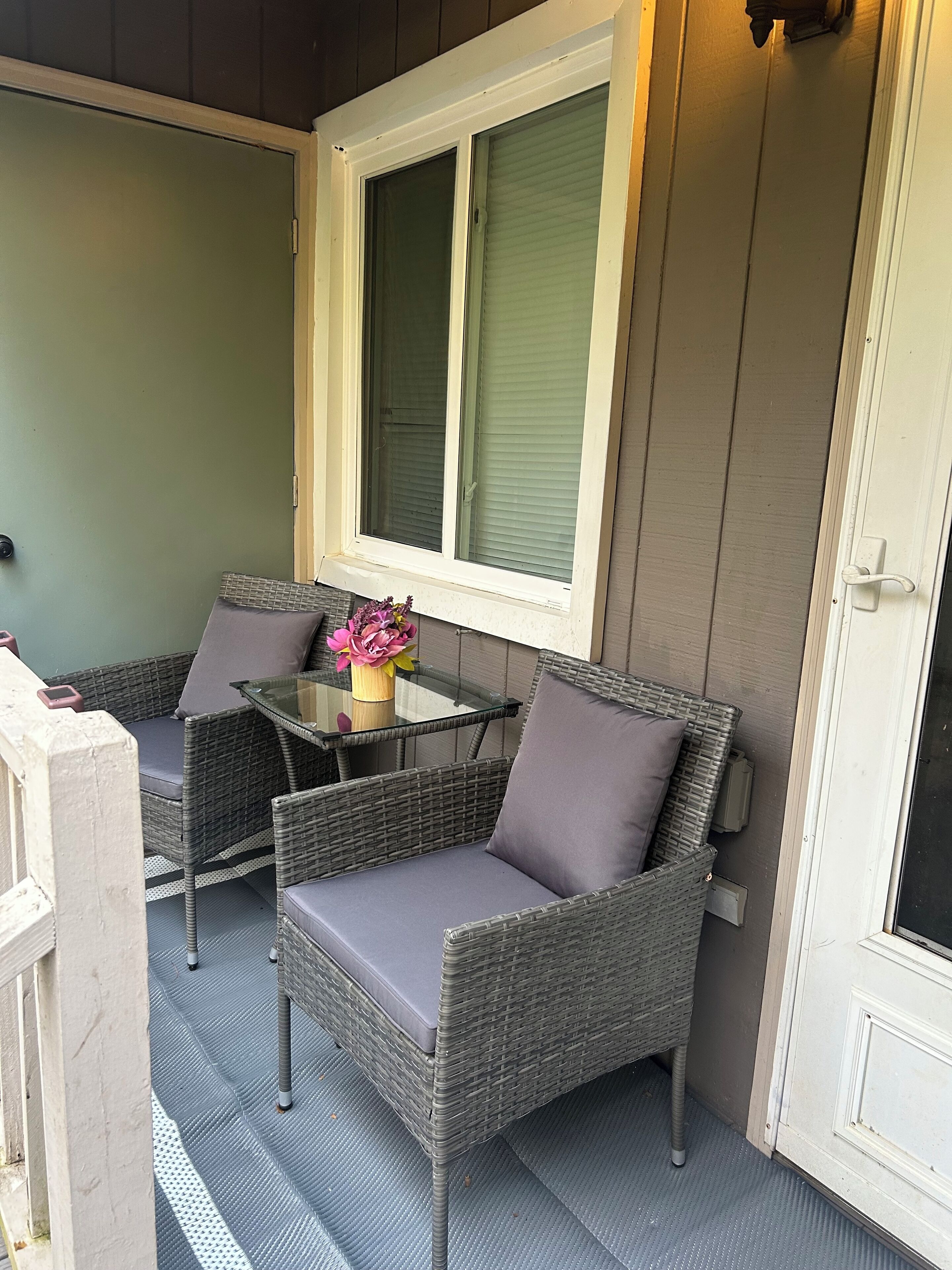 Ein kleiner Sitzbereich im Freien mit Korbstühlen und einem Glastisch, ein Fenster mit Rollläden und eine Tür mit einem Insektenschutzgitter.