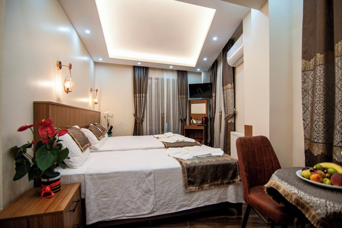 A hotel room with two beds, a dining table with fruit, a TV, and a large window with curtains.