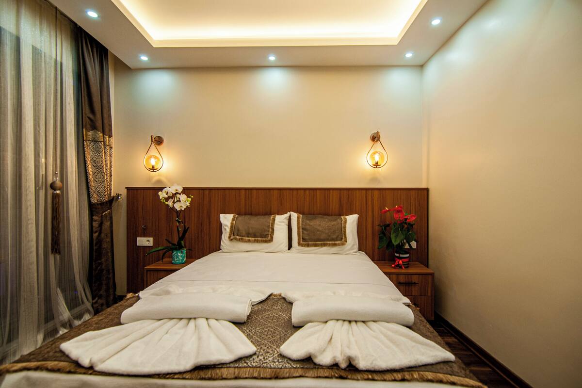 A hotel room with a neatly made bed, two bedside tables with lamps, and a large window with curtains.