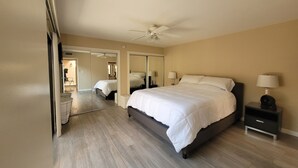 A bedroom with a large bed, a ceiling fan, a nightstand with a lamp, and a mirror on the wall.