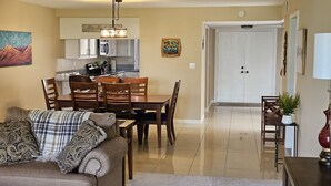 A living room with a sofa, a dining table with chairs, a kitchen area with cabinets and appliances, and a hallway leading to a door.