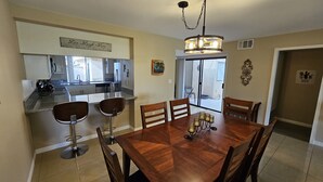 A dining area with a wooden table and chairs, a chandelier, and a kitchen in the background.