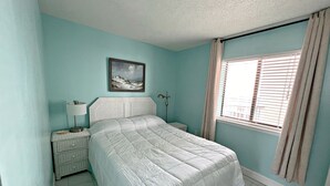 Ein Schlafzimmer mit Bett, Nachttisch, Lampe, einem Bild an der Wand und einem Fenster mit Vorhängen.